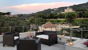 Una terraza con mobiliario de mimbre y velas, con vistas a un paisaje urbano de edificios y árboles.