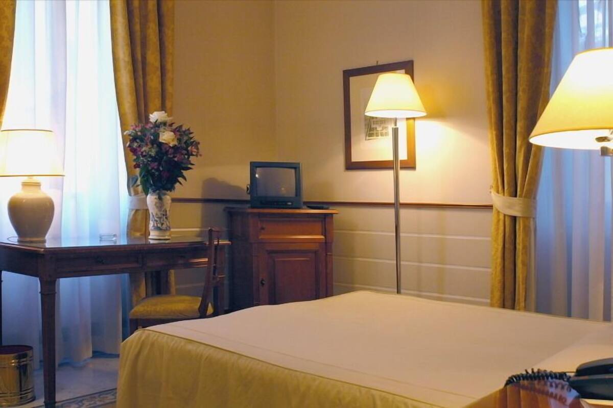 A hotel room with a bed, a desk with a lamp, a vase with flowers, a framed picture on the wall, and curtains.