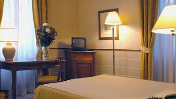 A hotel room with a bed, a desk with a lamp, a vase with flowers, a framed picture on the wall, and curtains.