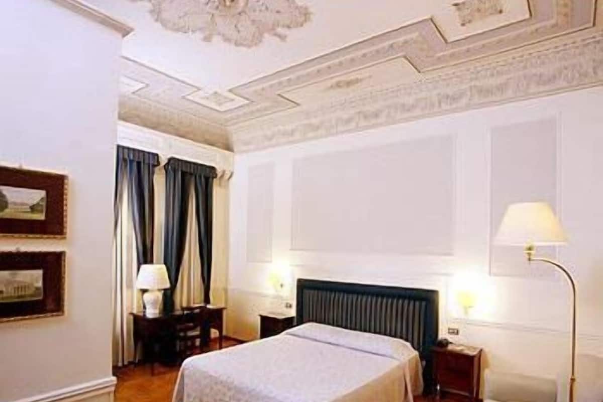 A hotel room with a large bed, two bedside tables with lamps, a chair, and a ceiling with decorative molding.