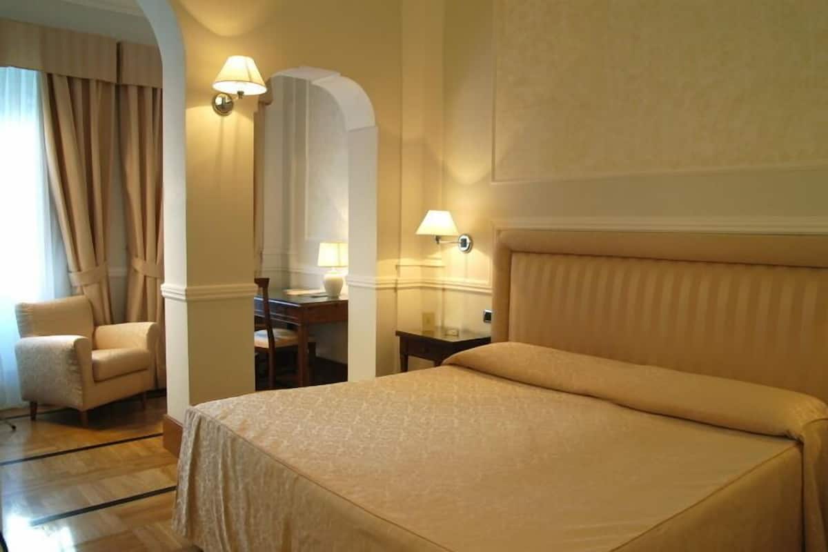 A hotel room with a large bed, a chair, a desk, and two wall-mounted lamps.