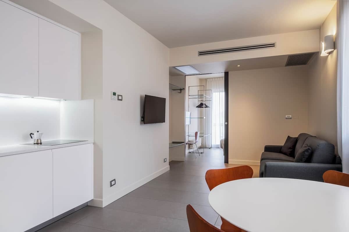 A modern kitchen with white cabinets, a coffee machine, and a flat-screen TV. There is a dining area with a white table and wooden chairs.