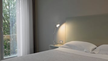 Una camera d'albergo con un letto, una lampada da comodino, una finestra con tende e vista sugli alberi.