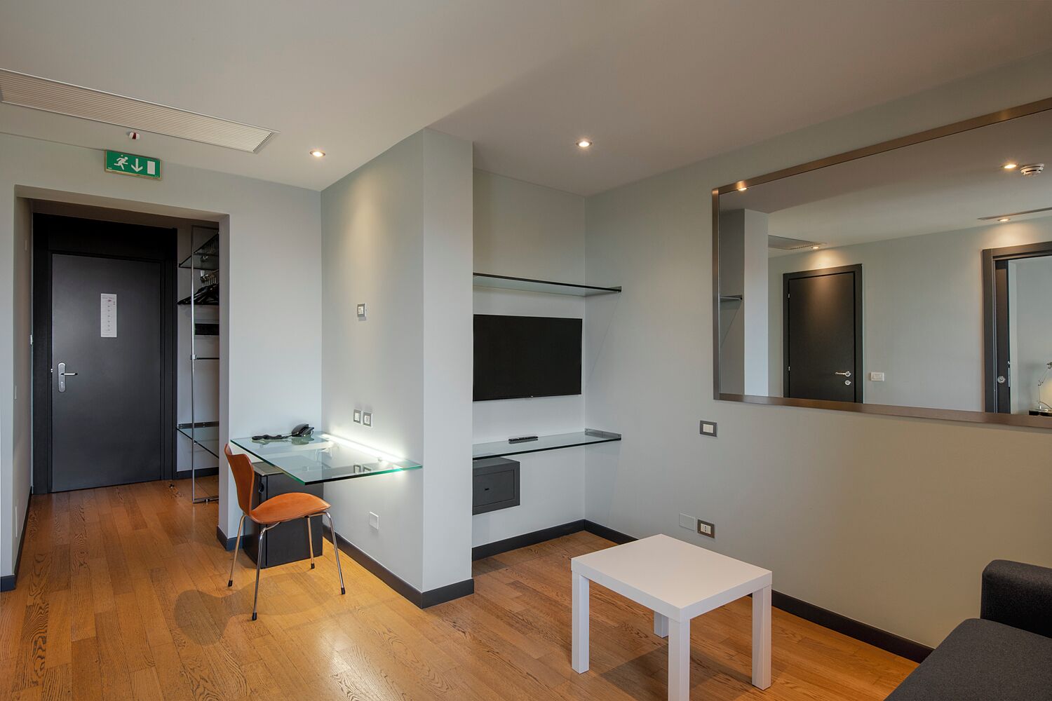 A modern hotel room with a glass desk, a small white table, a mirror, and a wardrobe.