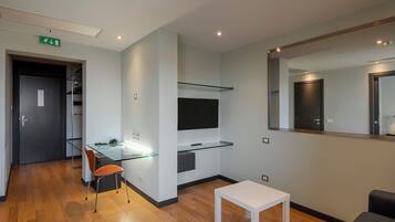 A modern hotel room with a glass desk, a small white table, a mirror, and a wardrobe.