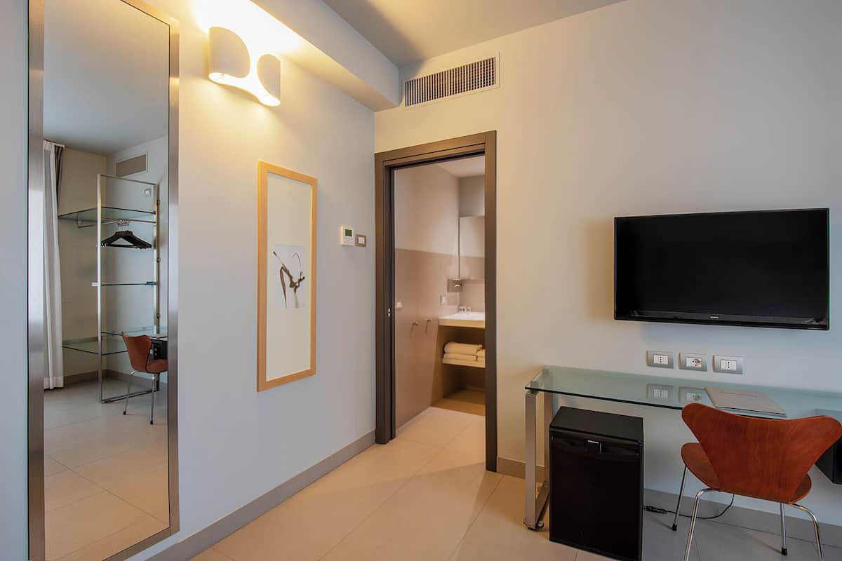 A modern hotel room with a glass desk, a flat-screen TV, a mirror, and a framed picture on the wall.