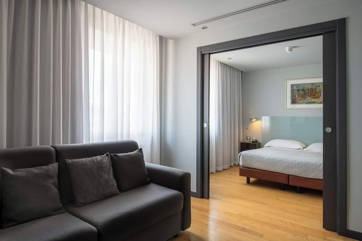 A modern hotel room with a grey sofa, a bed with a patterned duvet, and a painting on the wall.