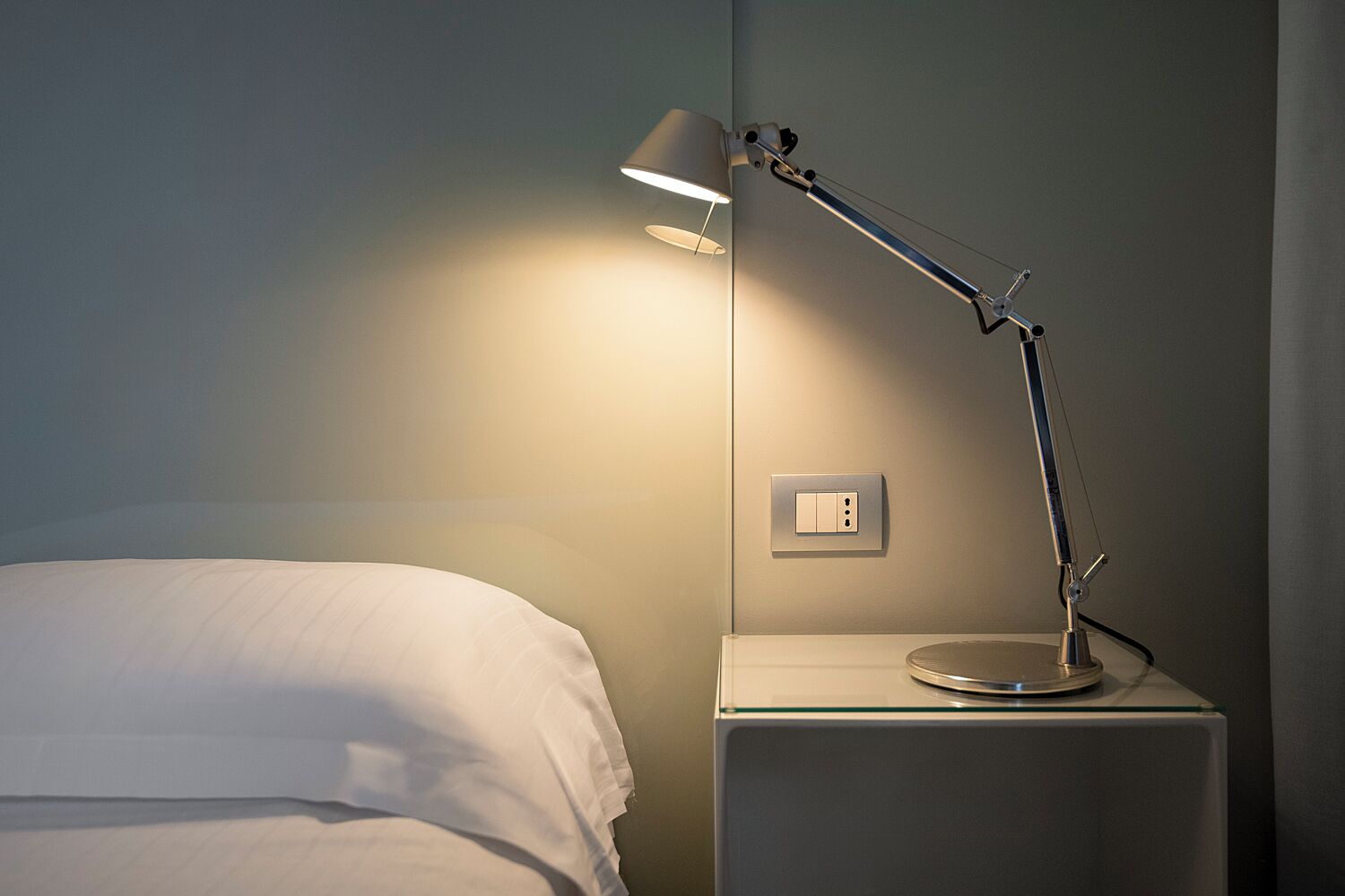 Una cama bien hecha con un almohadón blanco y una mesita de noche con una lámpara y un enchufe.