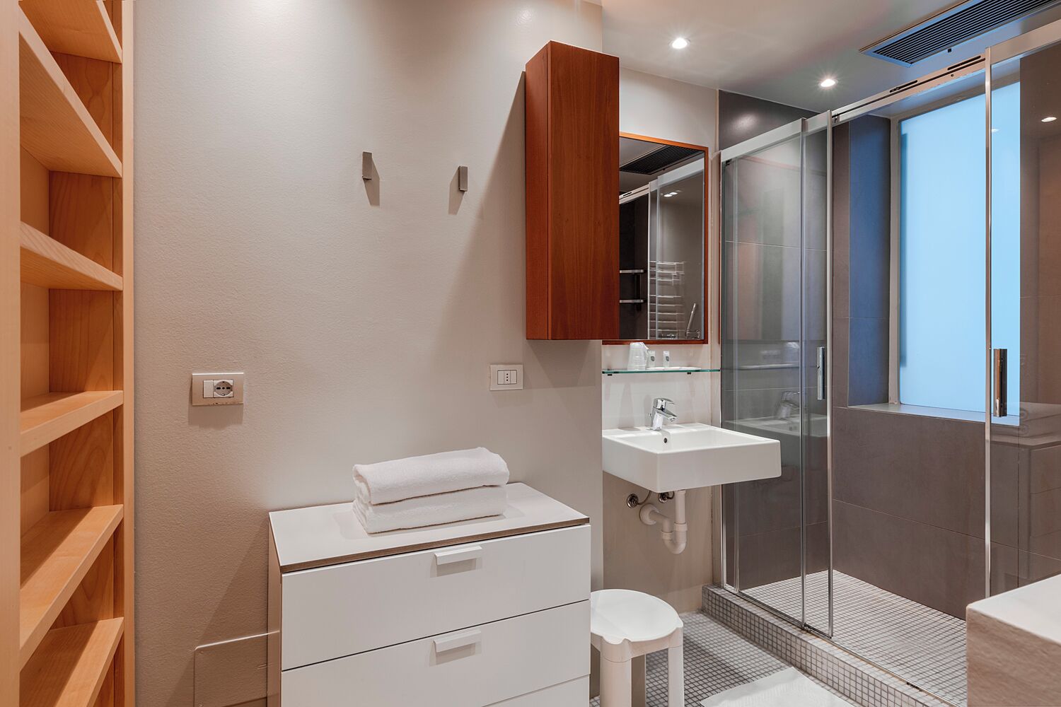 Un baño moderno con un lavamanos blanco, estantes de madera, un espejo y una cabina de ducha de vidrio.