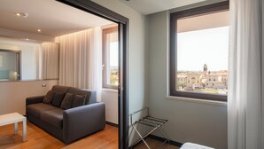 A modern bedroom with a grey sofa, a small white table, a metal drying rack, and a large window with a city view. - Hotel Verona (Verona)
