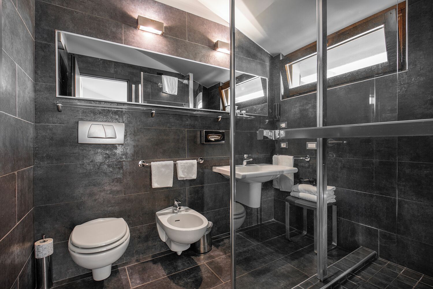 A modern bathroom with dark tiles, a glass shower enclosure, a toilet, bidet, and a sink.