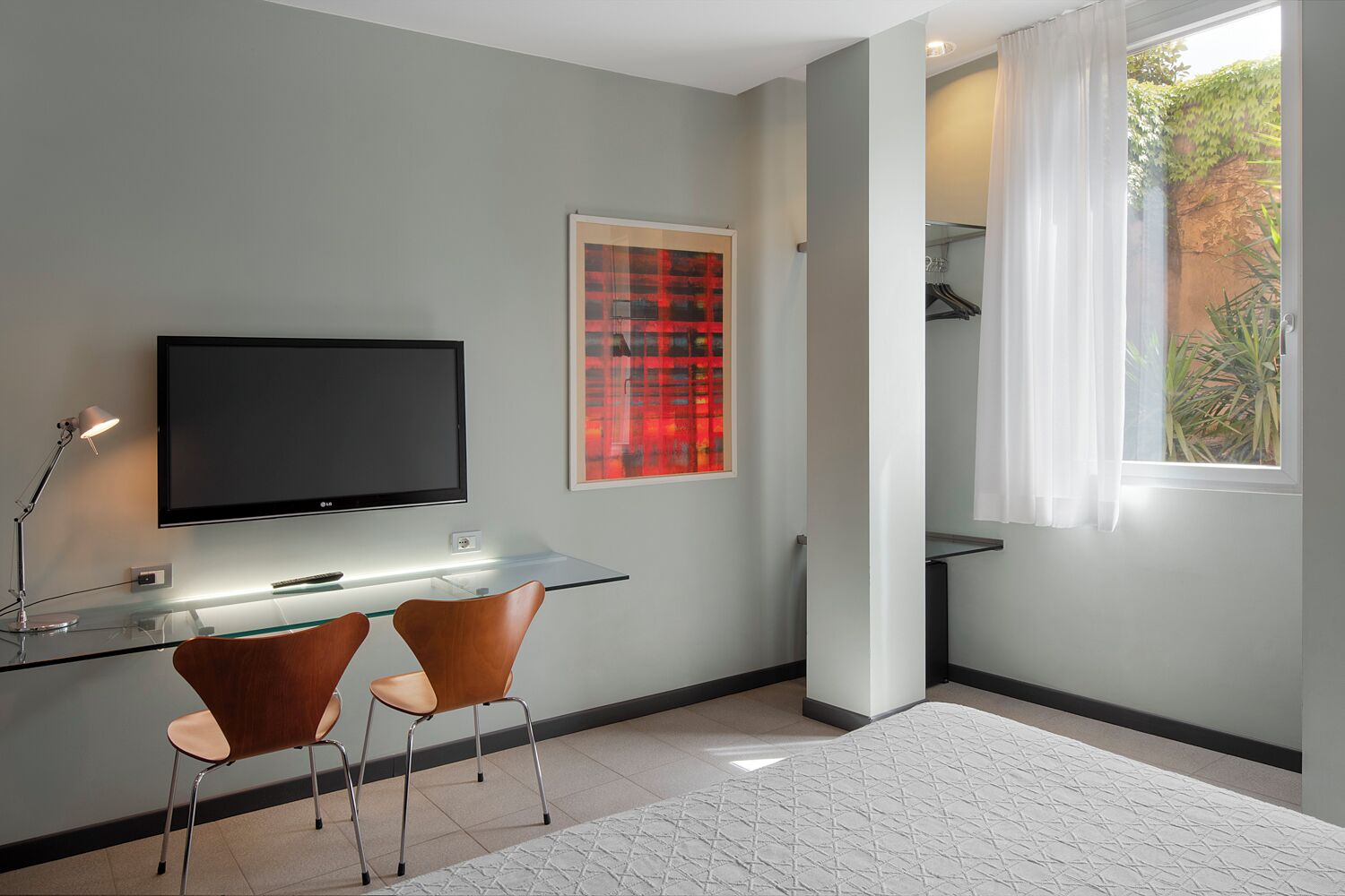 A modern room with a flat-screen TV, two chairs, a glass desk, a red artwork, and a window with curtains.