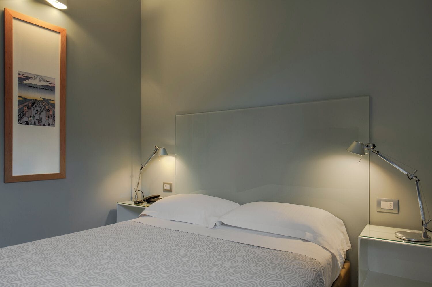 A neatly made bed with white bedding, two bedside lamps, and a framed picture of a mountain on the wall.