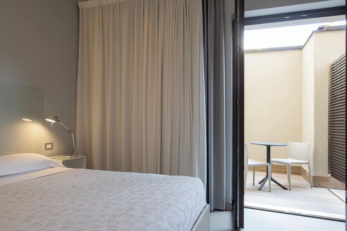 A hotel room with a bed, a bedside lamp, and a view of an outdoor seating area.