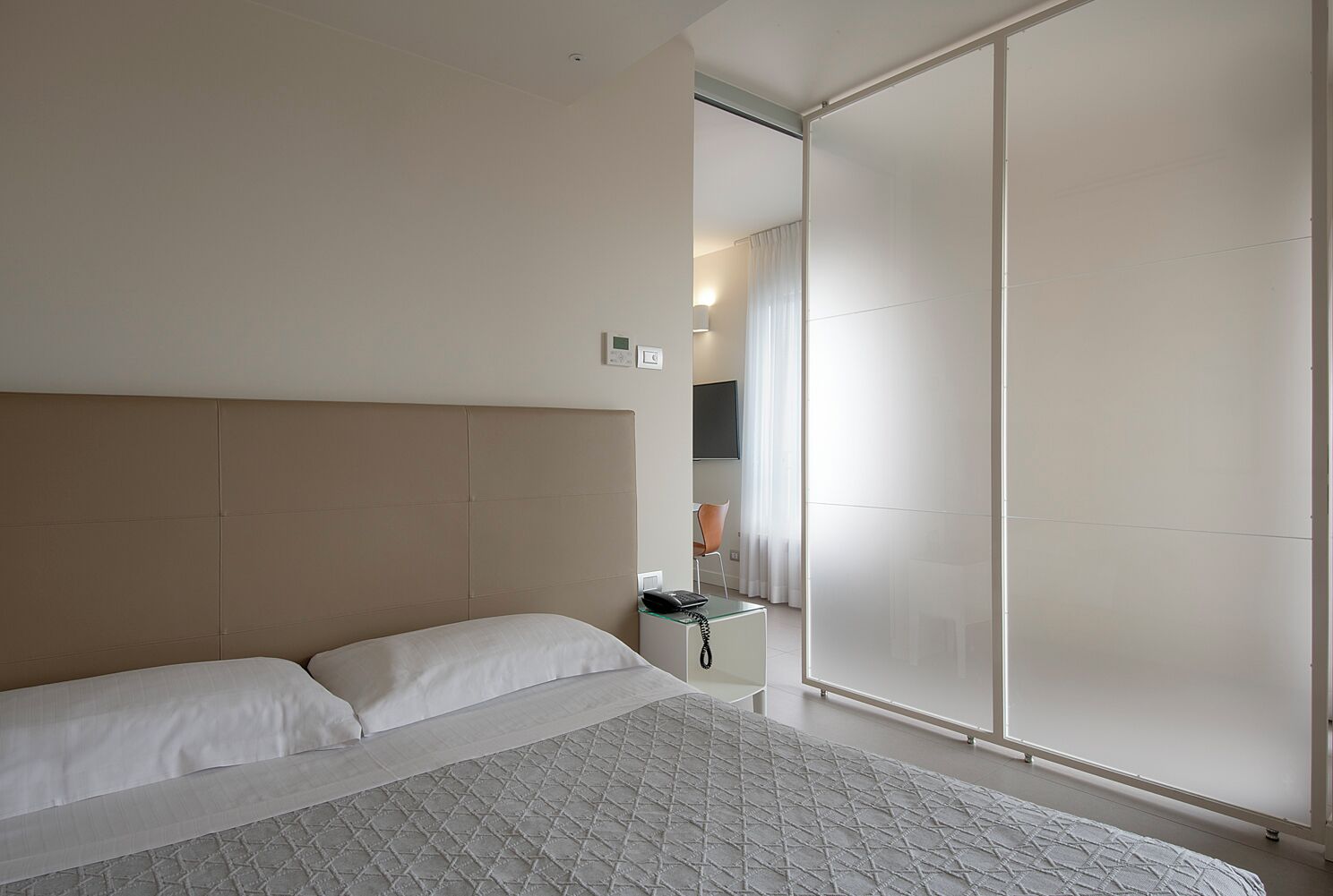 A modern hotel room with a large bed, a bedside table, a desk, and a sliding door leading to a bathroom.