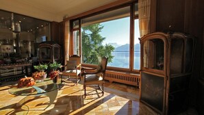 Quarto espaçoso com uma grande janela que oferece vista para um lago e montanhas. Há uma mesa de centro de vidro, duas poltronas estofadas e um armário de madeira.