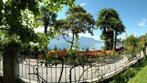 Um jardim bem cuidado com diversas árvores e arbustos, um caminho pavimentado e vista para o mar.