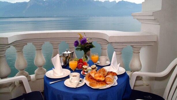 Mesa posta com itens de café da manhã, incluindo croissants, frutas e suco, com vista para um lago e montanhas ao fundo.