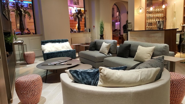 A modern living room with a grey sofa, a blue armchair, and a round coffee table. There is a bar area with glasses and bottles in the background. - Best Western Plus Nice Cosy Hôtel (Nice)