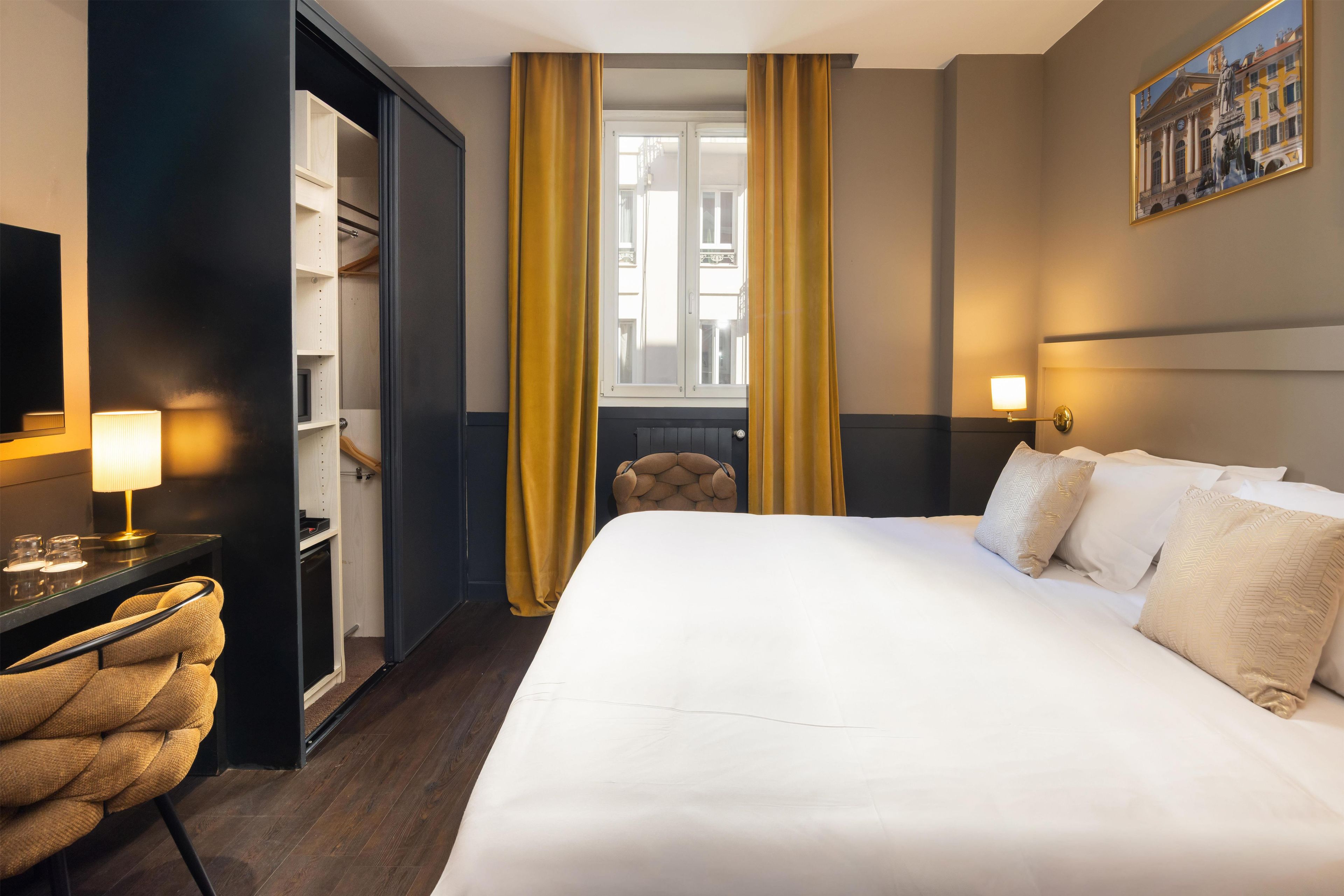 Habitación de hotel con cama, escritorio, silla, armario y ventana con cortinas.