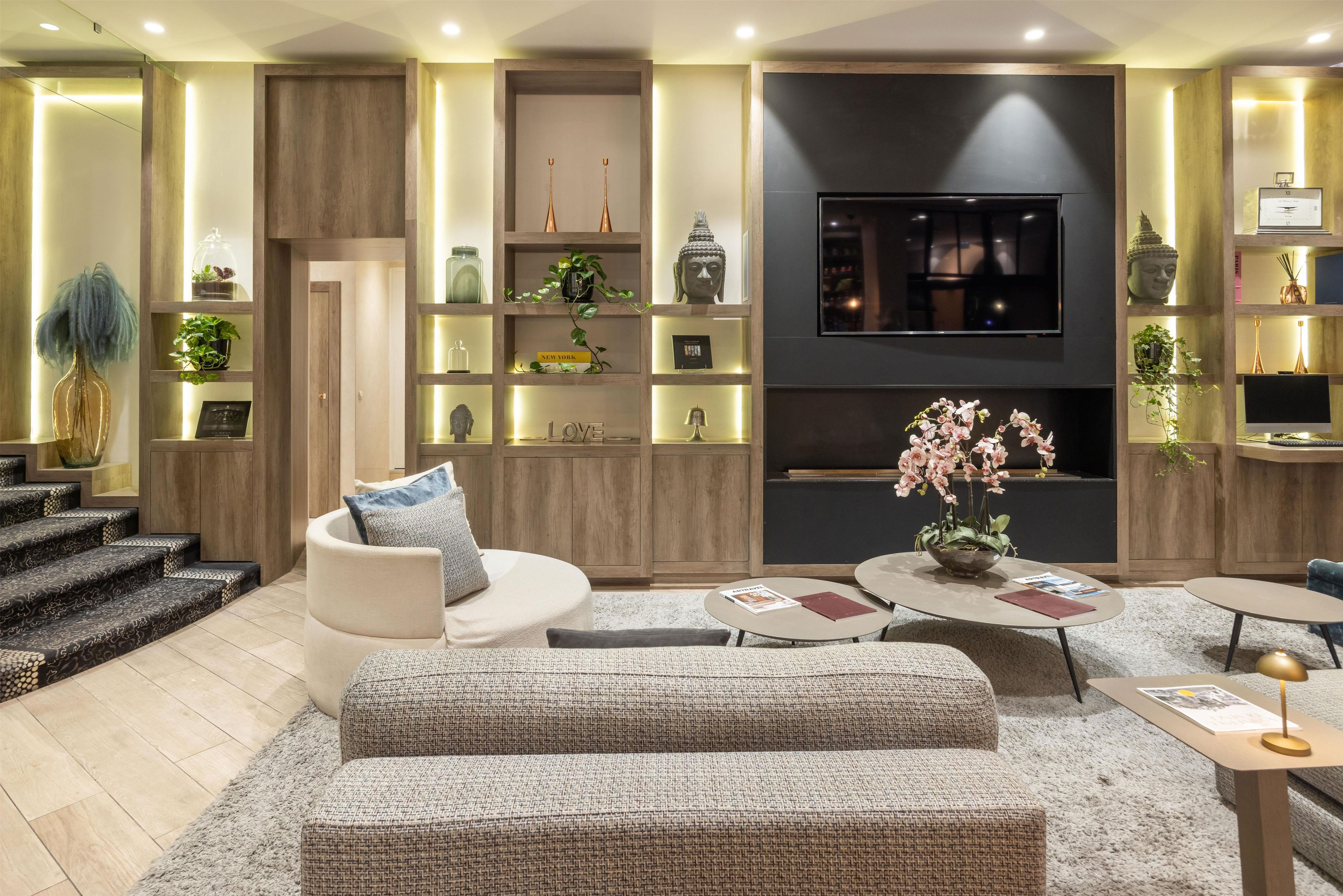 Una sala de estar moderna con sofá, mesa de centro y televisor. Se ven estantes con adornos y una escalera.