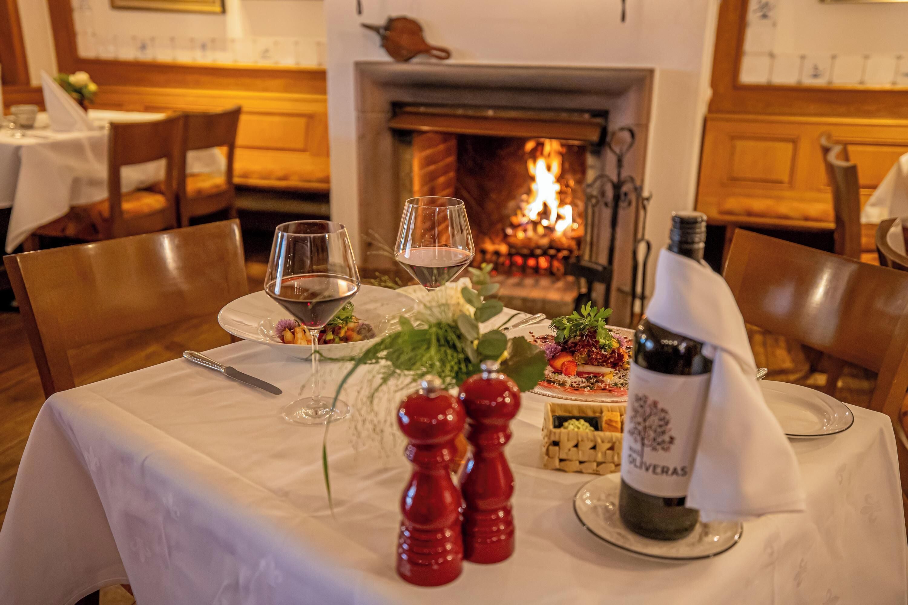 A dining table set with wine glasses, a bottle of wine, salt and pepper shakers, and a small fruit dish. A fireplace is visible in the background.