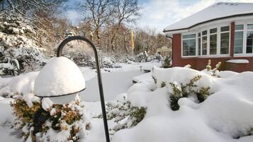 Un jardín cubierto de nieve, con un farol de metal y un edificio con ventanas grandes al fondo.