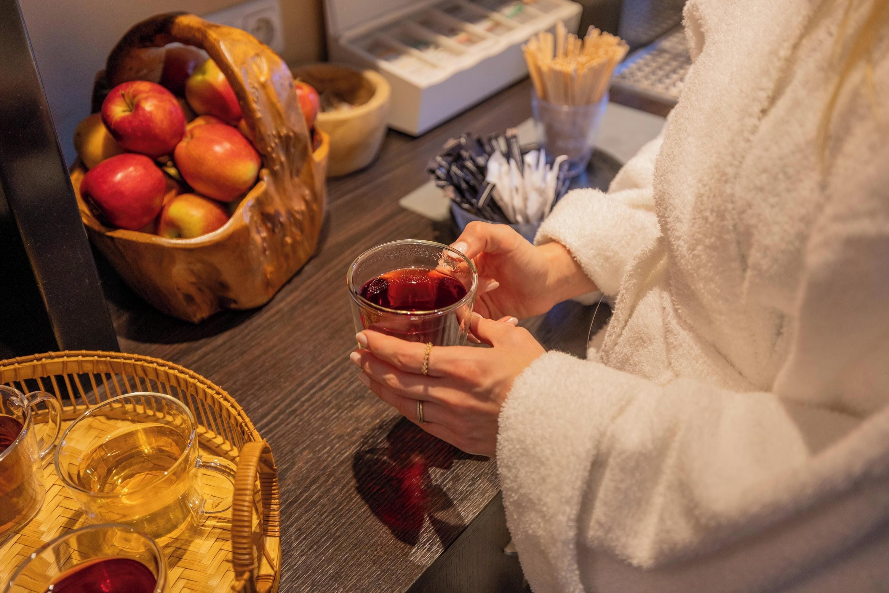 A person holding a glass of red drink, a basket of apples, and a tray with glasses on a wooden table.