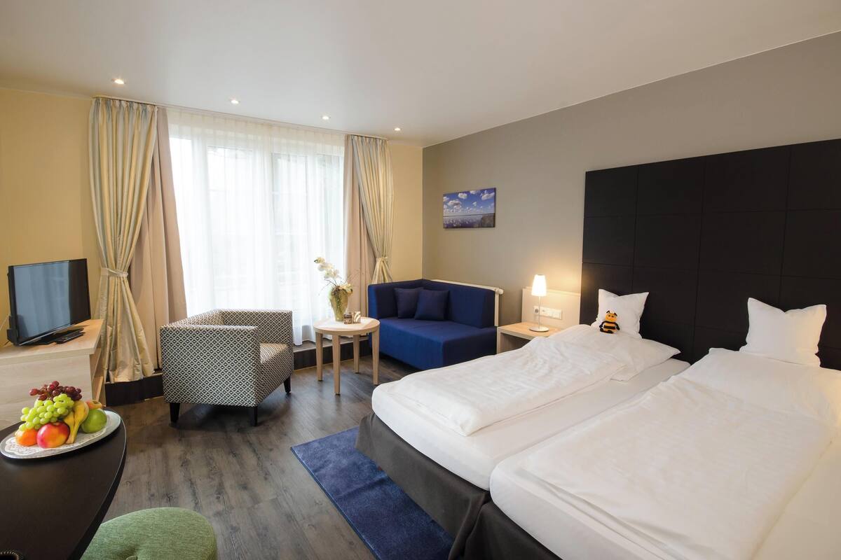 Hotellihuone, jossa on sänky, televisio, työpöytä tuolilla, sohva ja pieni pöytä, jonka päällä on maljakko täynnä kukkia.