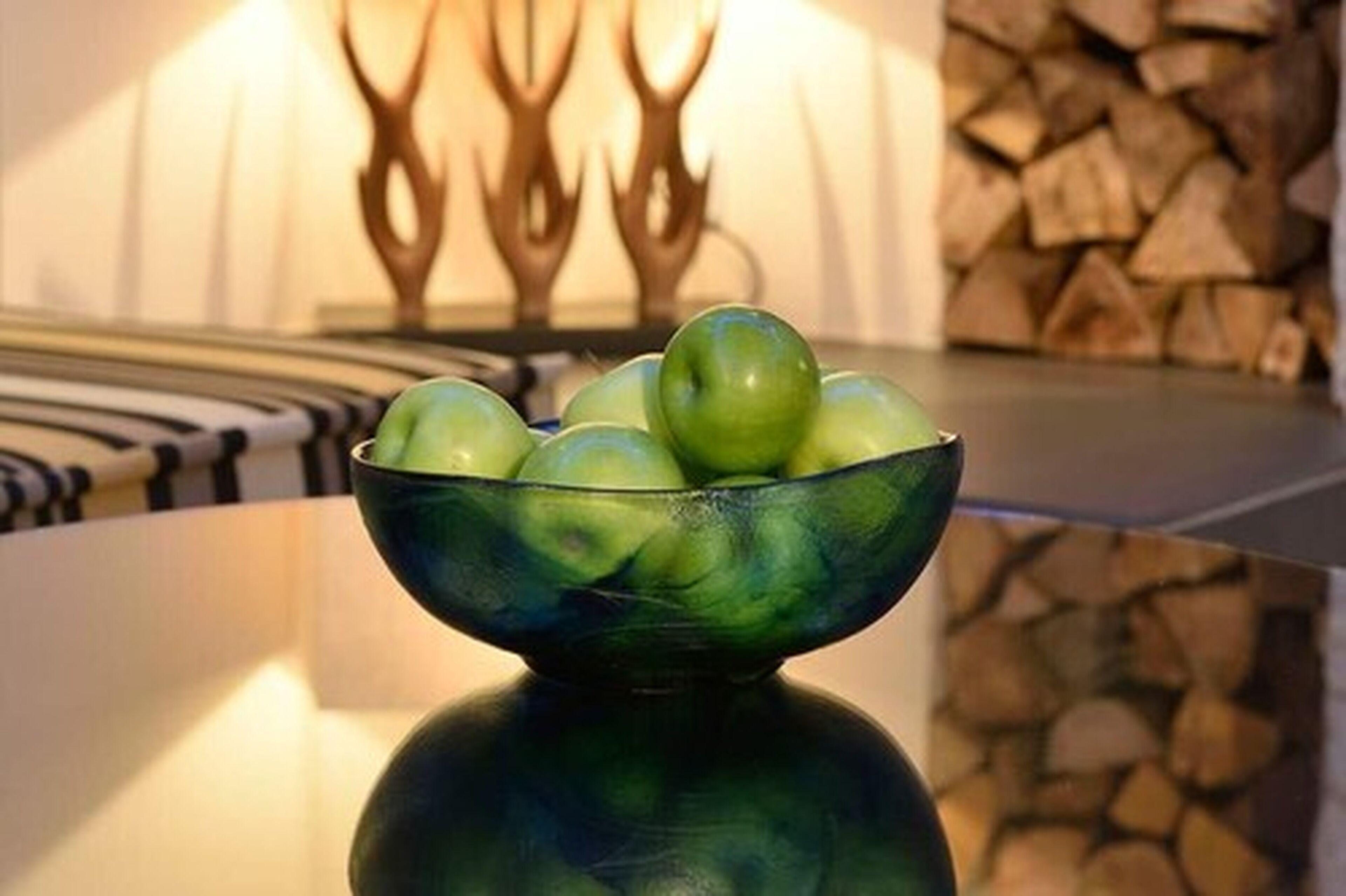 A bowl of green apples on a reflective surface with a blurred background featuring a decorative wall and stacked logs.