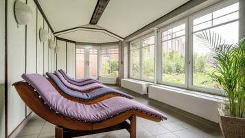 A spa room with multiple massage tables aligned against the wall, large windows, and a potted plant.