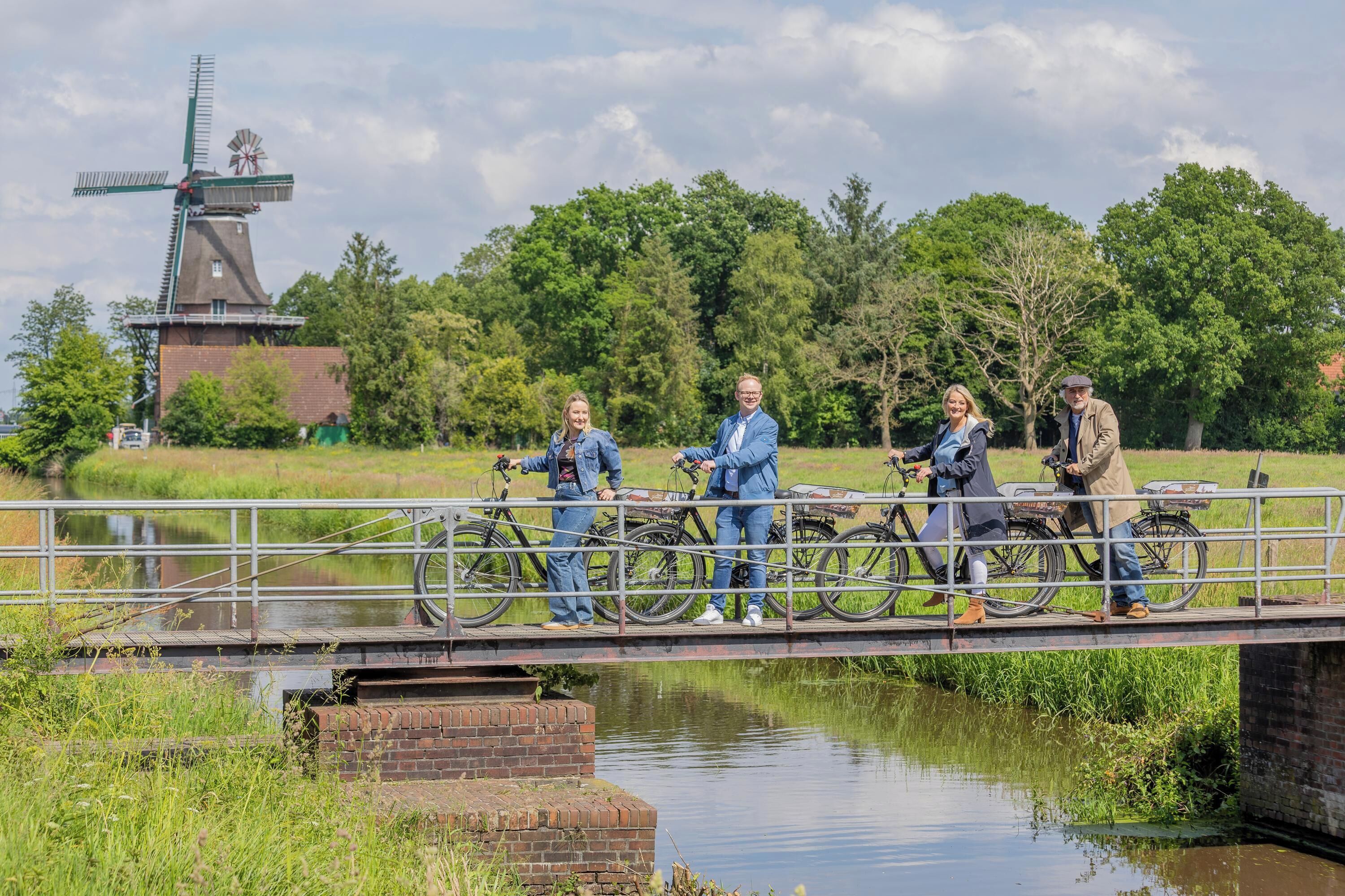 A group of people on a bridge with bicycles, a windmill in the background, and a body of water below.