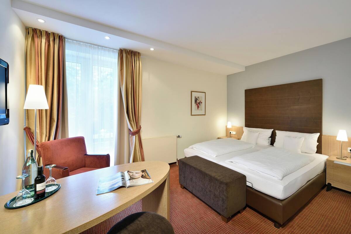 Hotellihuone, jossa on suuri sänky, työpöytä, jossa on lehti, tuoli ja ikkuna, jossa on verhot.