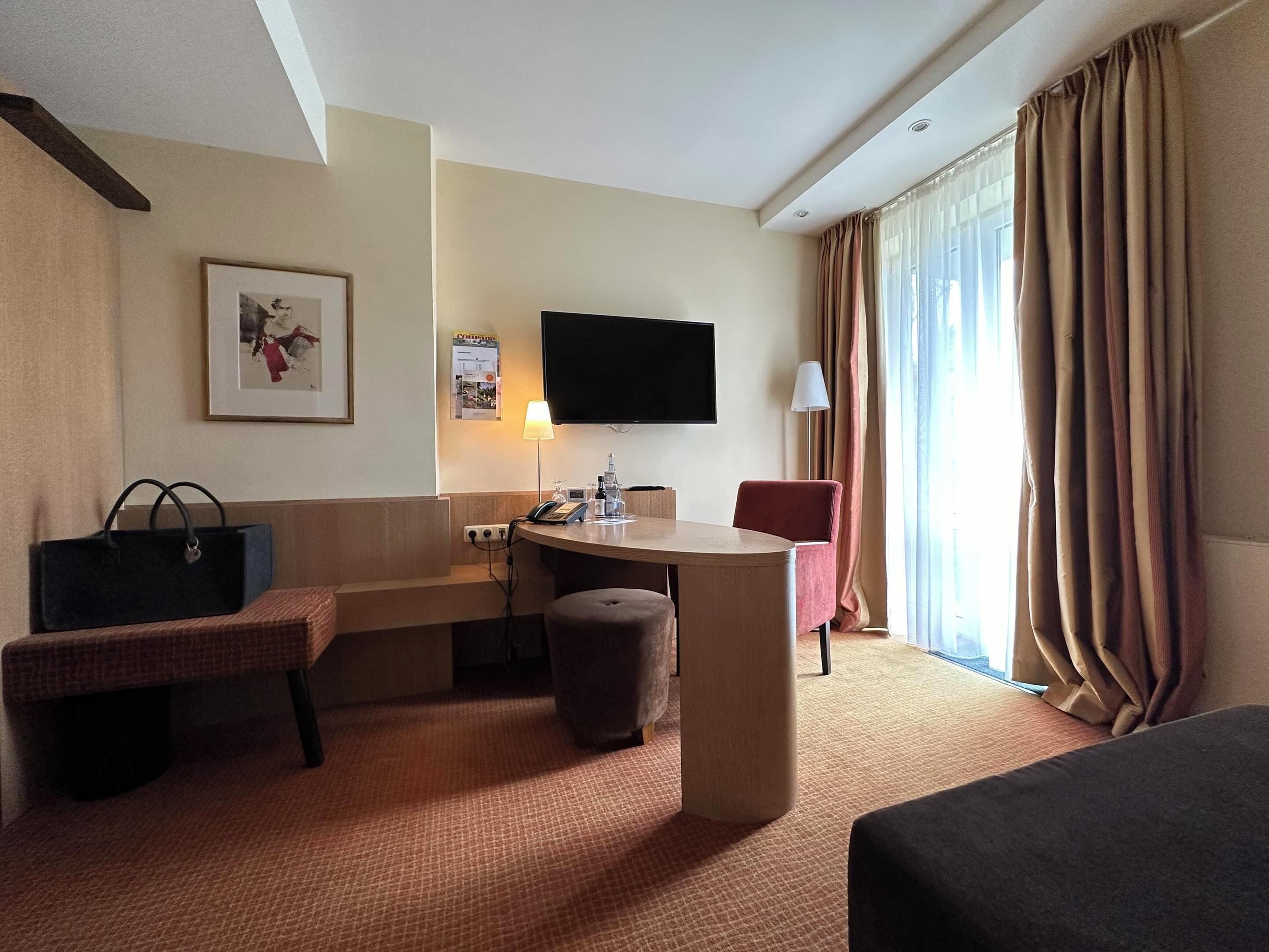 Ein Hotelzimmer mit Bett, Schreibtisch mit Lampe, Stuhl, Fernseher und einem Fenster mit Vorhängen.