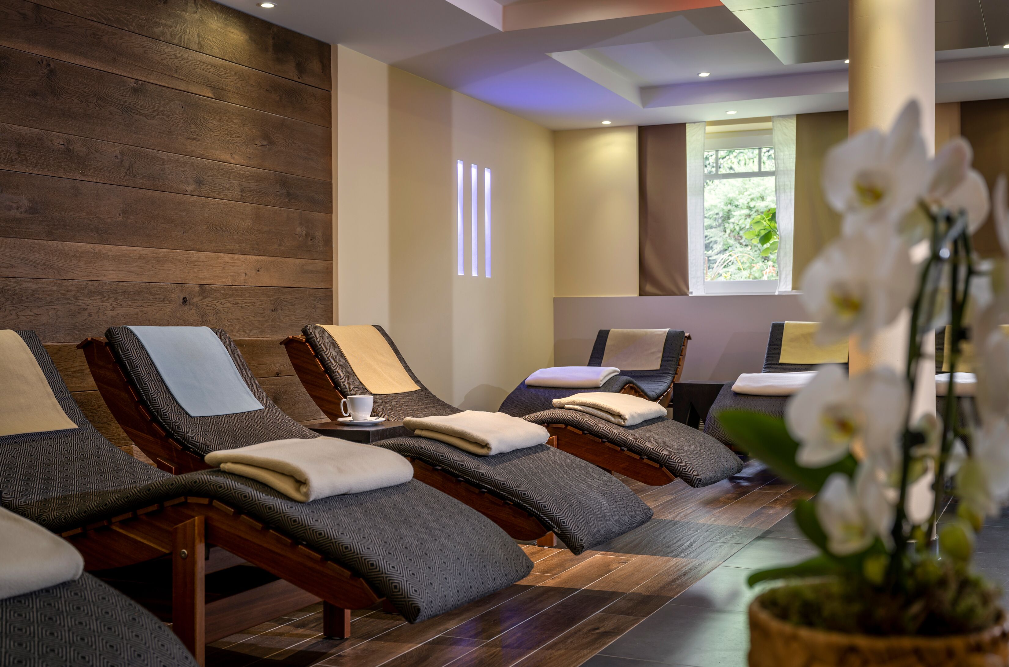 Um espaço de spa com paredes de madeira, várias cadeiras de massagem, uma orquídea em vaso e uma janela com vista para o verde.