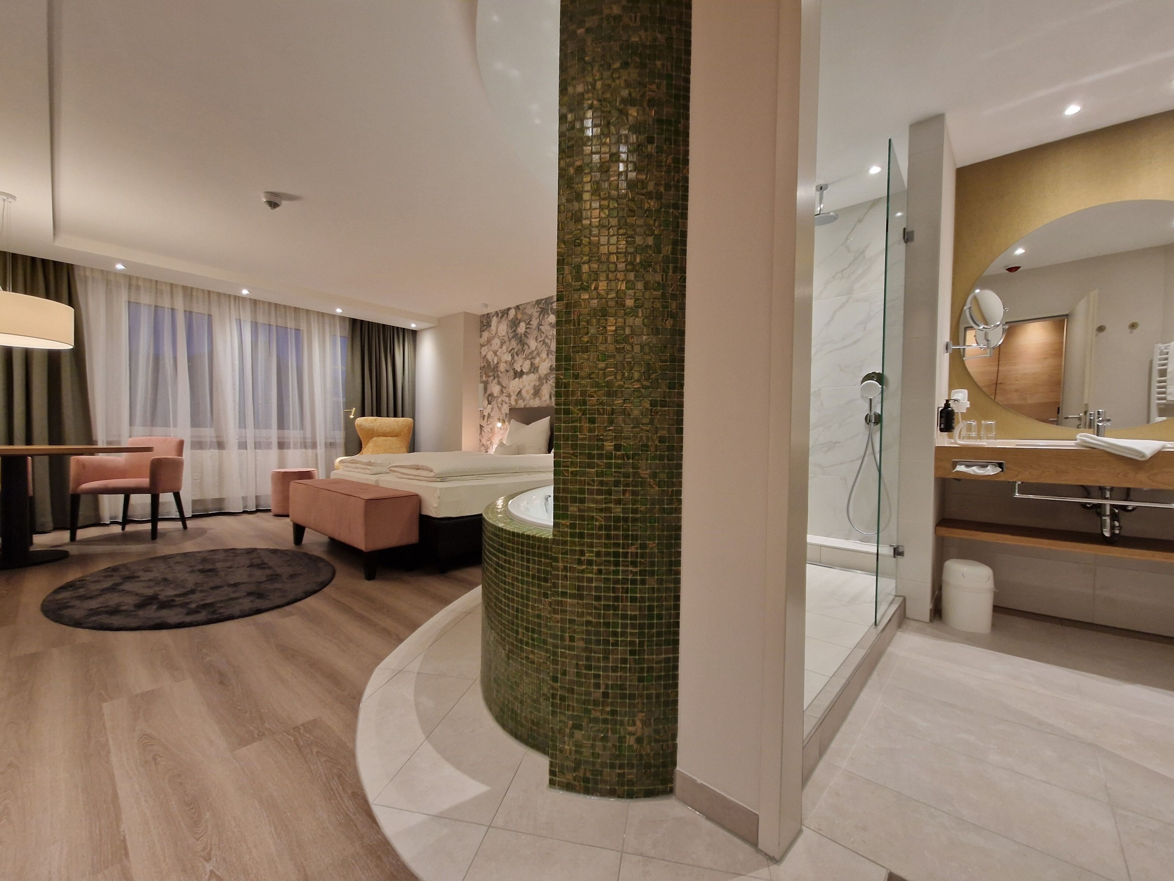 A modern hotel room with a bed, a sitting area with a chair and a table, a bathroom with a bathtub and a shower, and a large mirror.