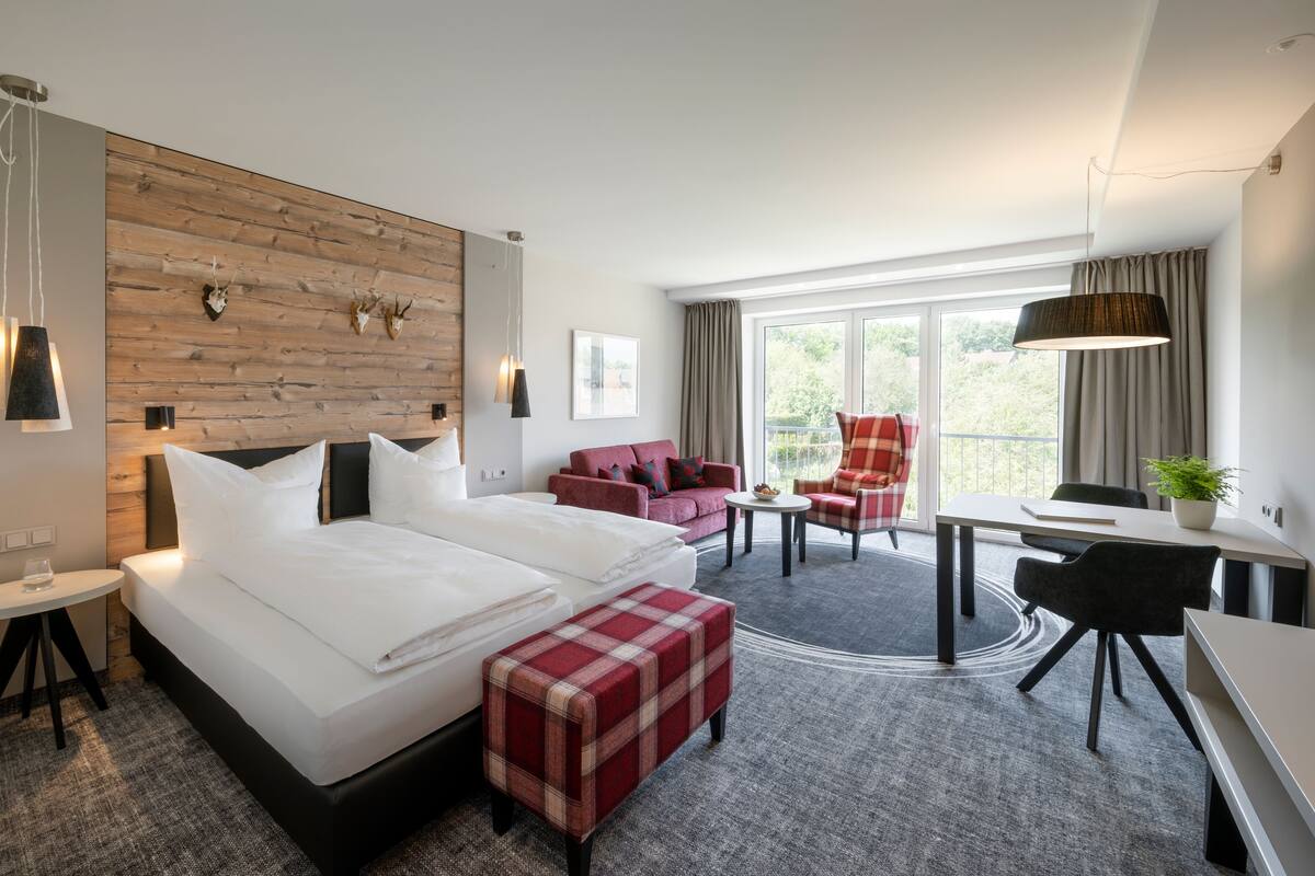 Hotellihuoneessa on suuri sänky, punainen sohva, työpöytä ja tuoli. Huoneessa on ikkuna, jossa on verhot, ja ikkunasta avautuu näkymä vehreään maisemaan.
