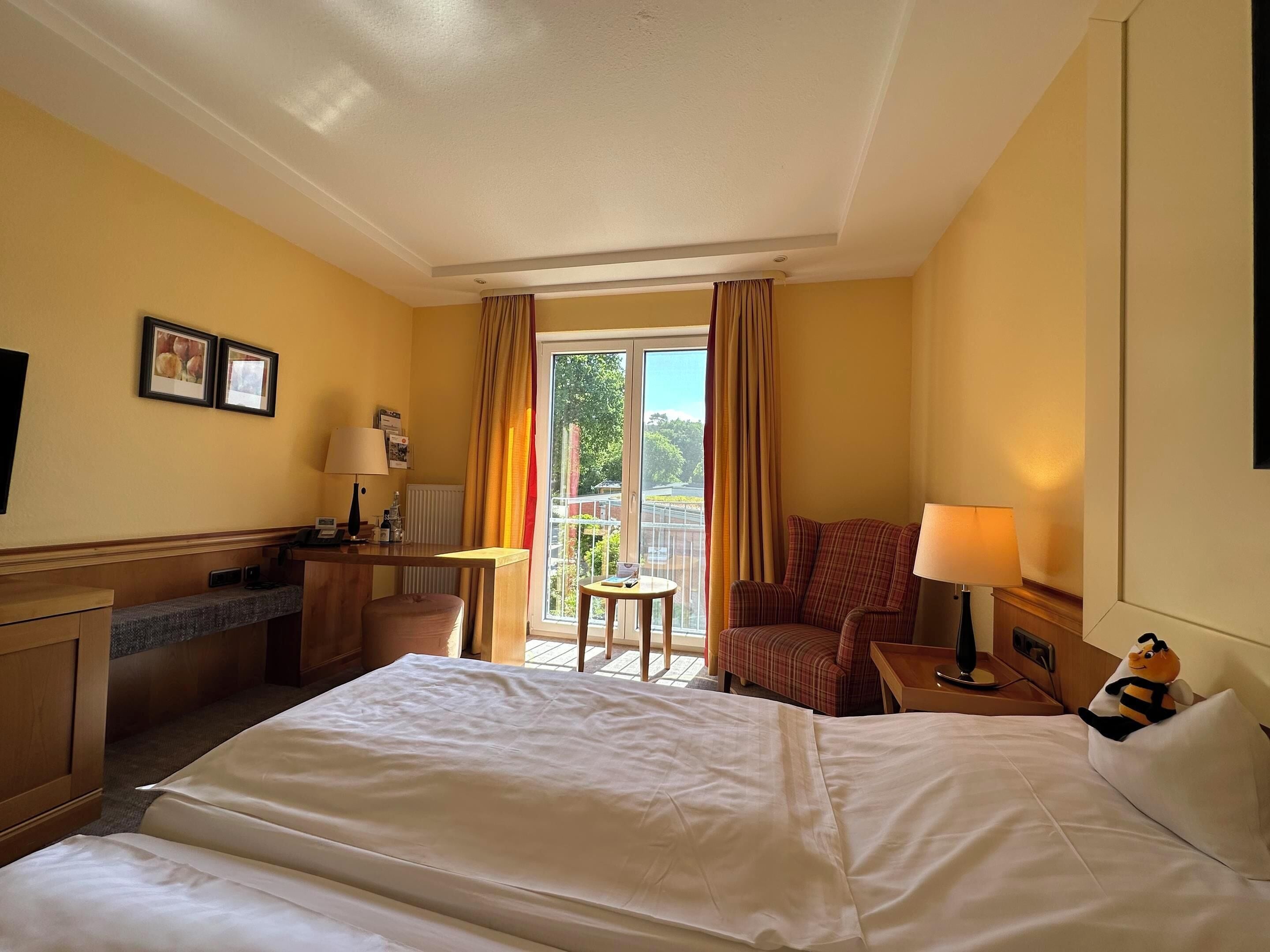 Ein Hotelzimmer mit einem Bett, einem Schreibtisch, einem Sessel und einem kleinen Tisch. Ein Fenster mit Vorhängen bietet Blick auf eine grüne Umgebung.