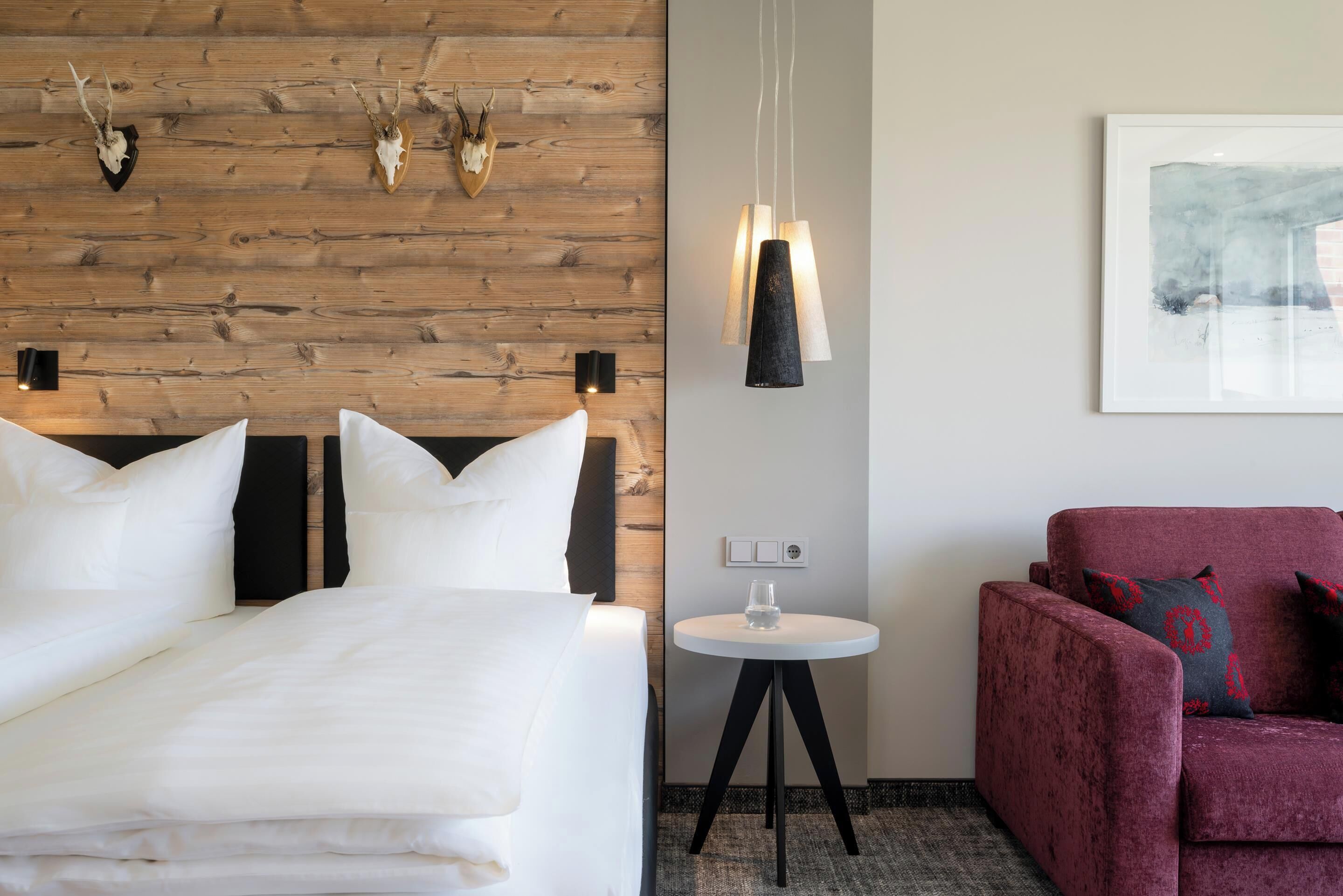 A hotel room with a wooden wall, a bed with white bedding, a red armchair, a small round table with a glass, and a modern pendant light.