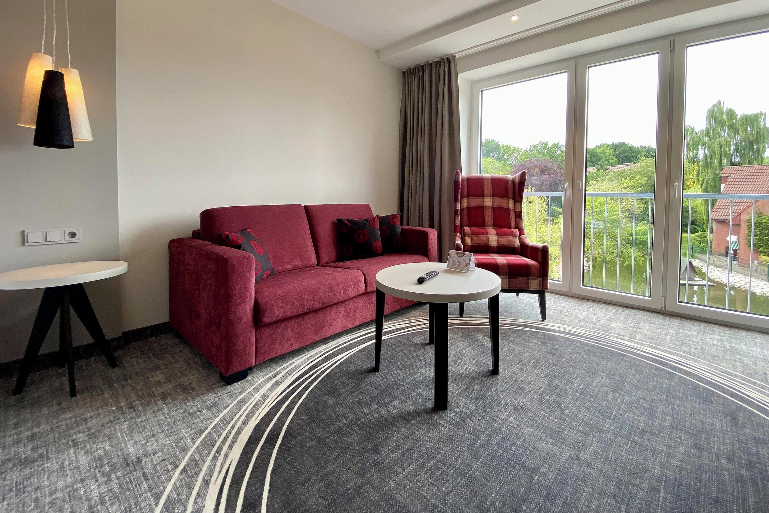 A modern hotel room with a red sofa, a plaid armchair, a small round table, and a large window offering a view of greenery and buildings.