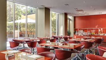 A modern restaurant with red chairs and tables, large windows, and a kitchen area.