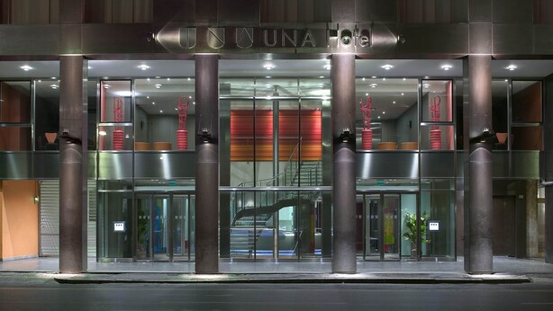 Une entrée d’hôtel moderne avec une façade en verre, un intérieur visible et un escalier menant à l’étage supérieur.