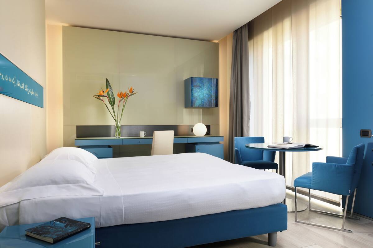 Een hotelkamer met een bed, een bureau, een stoel, een klein tafeltje en een vaas met bloemen.