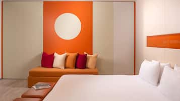 A modern hotel room with a large bed, a sofa with multiple pillows, a small table with a magazine, and a wall with a large circular design.