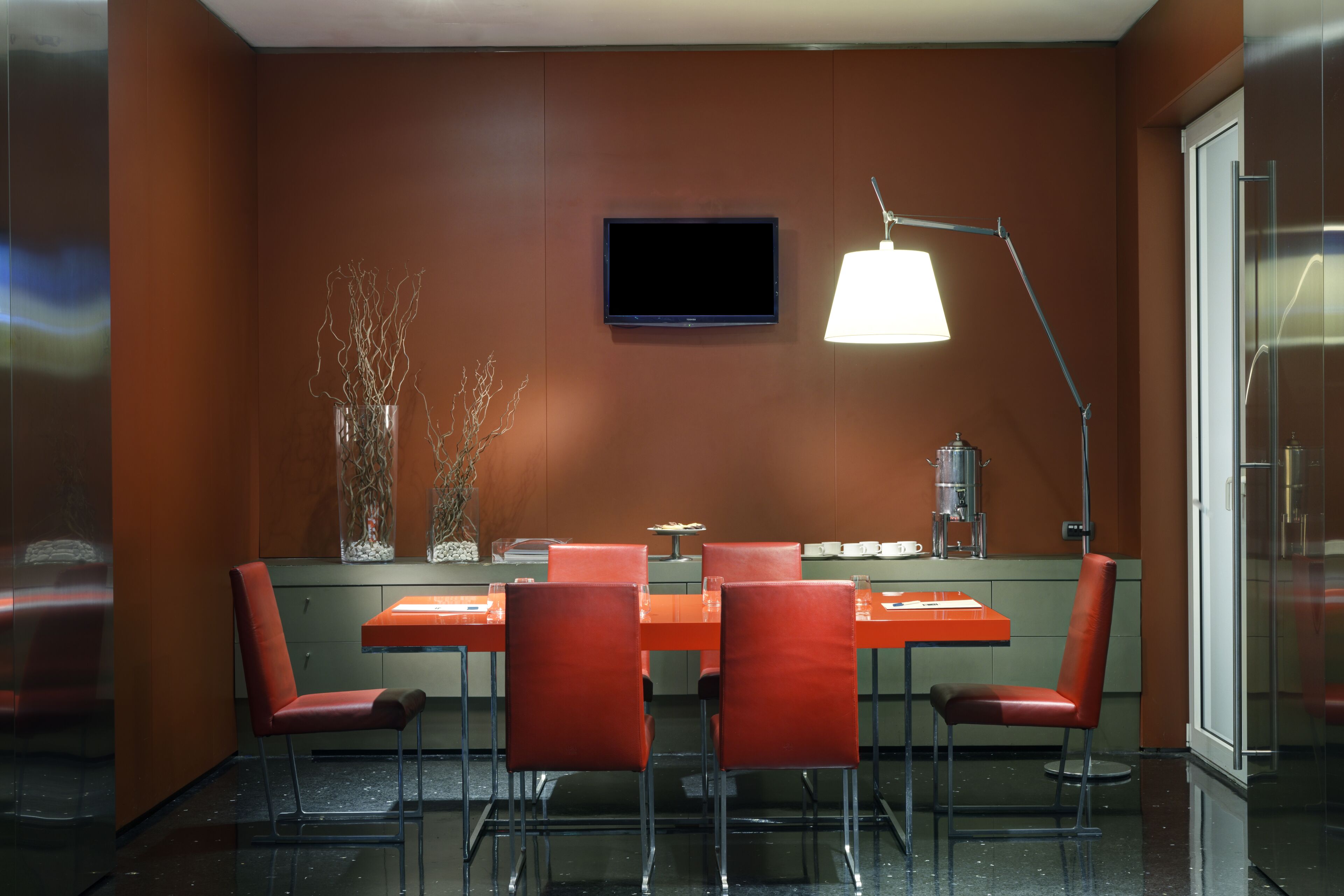 Uma sala de conferências moderna com uma mesa comprida, quatro cadeiras, uma TV de tela plana e uma luminária pendente.