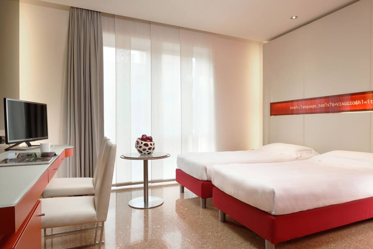 Een hotelkamer met een bed, een bureau met een computer, een stoel, een klein tafeltje met een schaal fruit, en een raam met gordijnen.