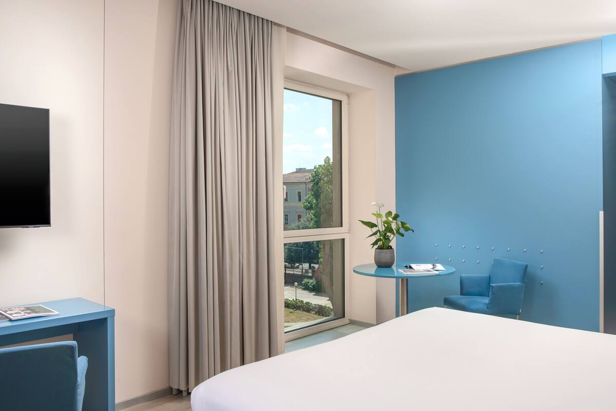 Een hotelkamer met een bed, bureau, stoel, televisie, raam met gordijnen en een potplant.