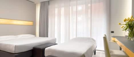 Habitación de hotel con dos camas, un escritorio, una silla, una mesita y un ventanal con cortinas traslúcidas.