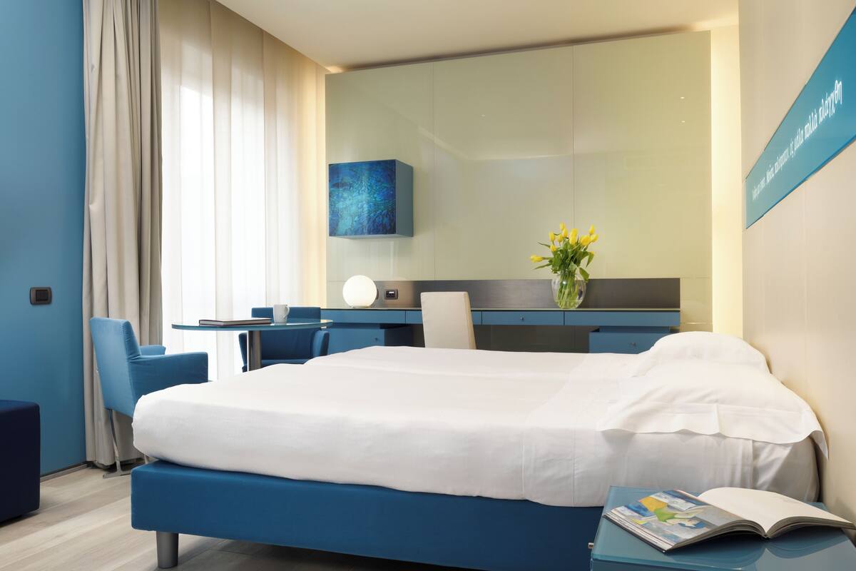 Een moderne hotelkamer met een groot bed, een bureau en een stoel. Op het bureau staat een vaas met bloemen en aan de muur een bord.