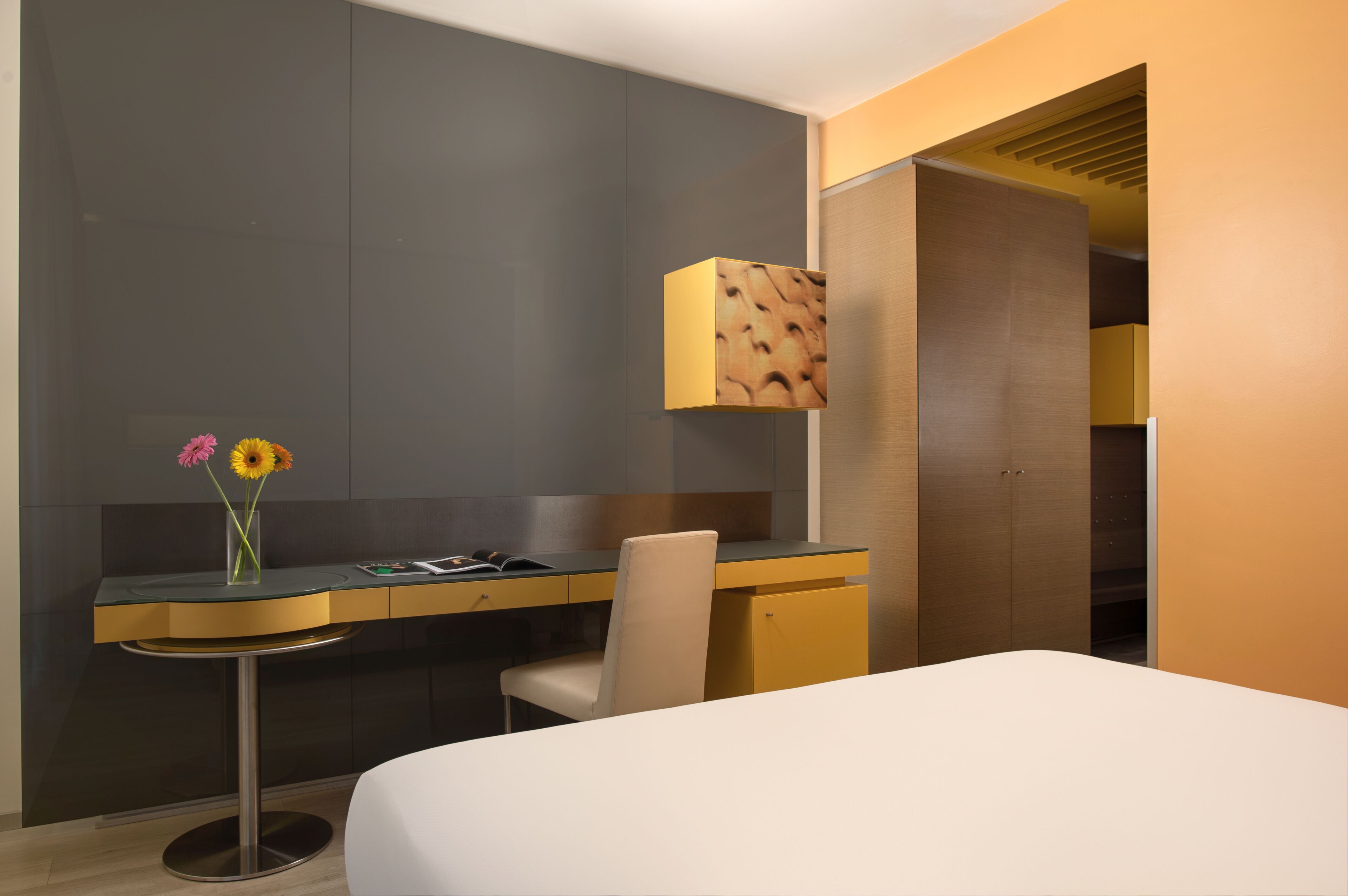 A modern hotel room with a bed, a desk with a chair, a vase with flowers, and a lamp.